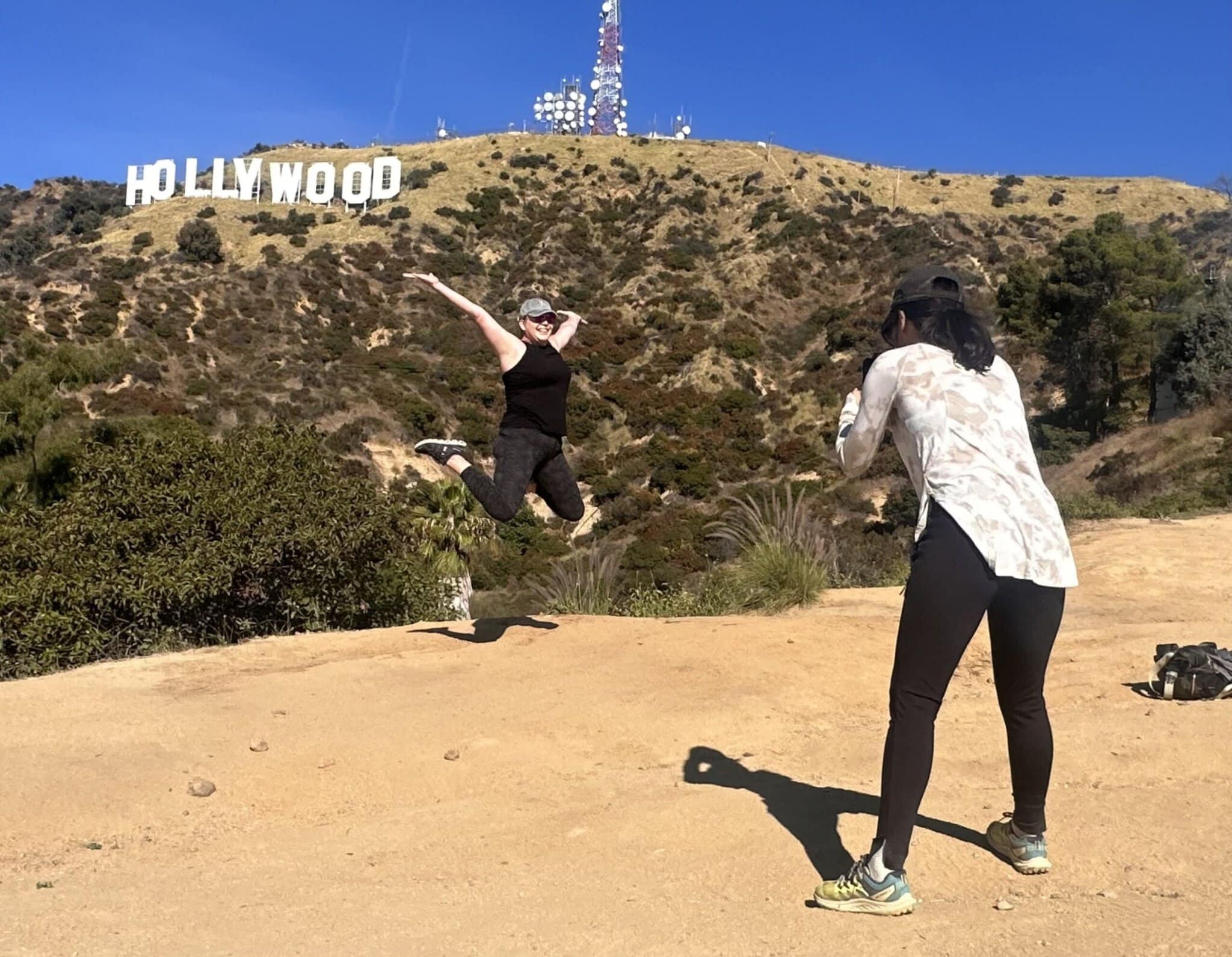 A girl takes a photo of another girl jumping in front of the Hollywood Sign on the Hollywood Sign Tour