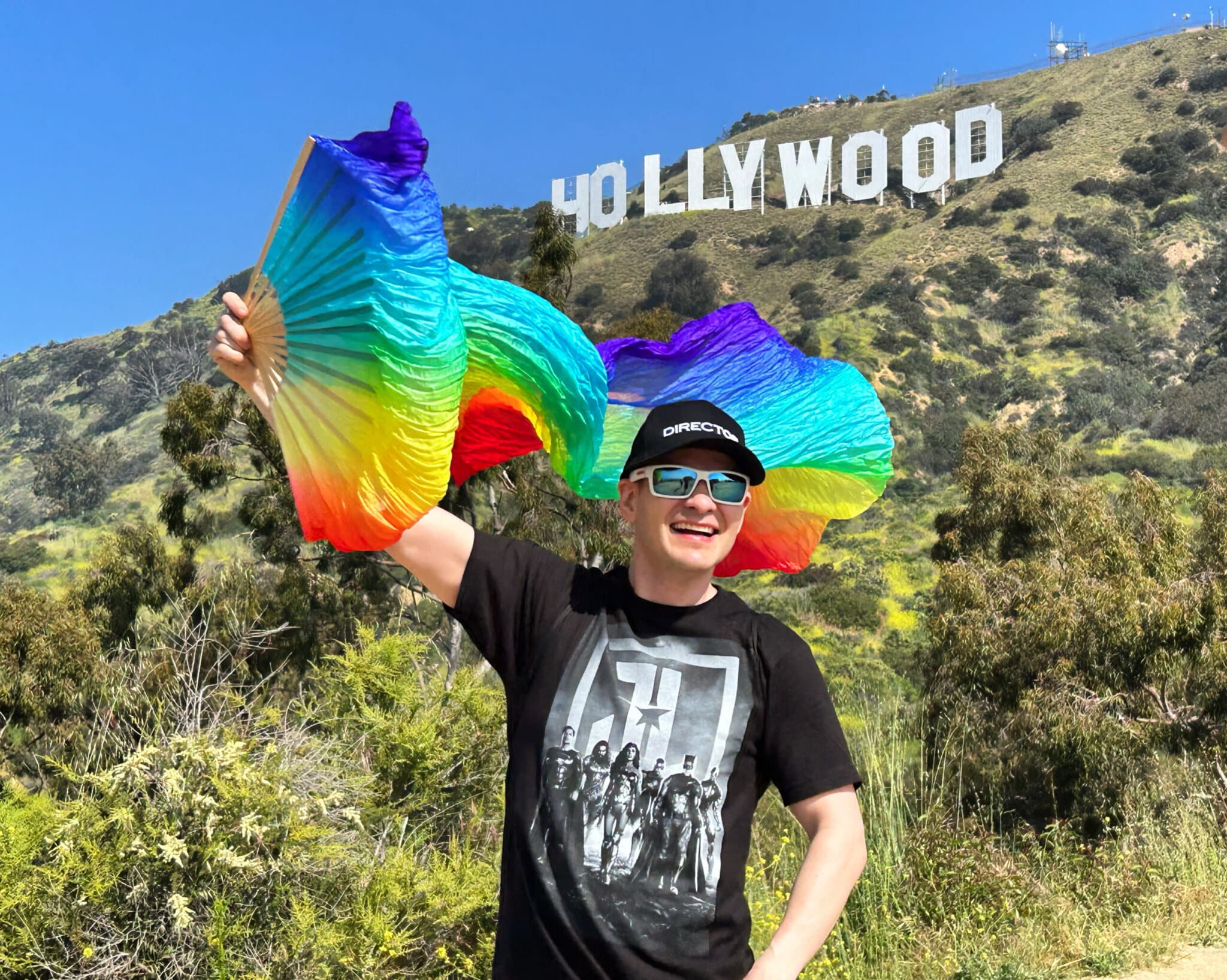 A man takes a photo holding a pride flag with the Hollywood Sign in the Background