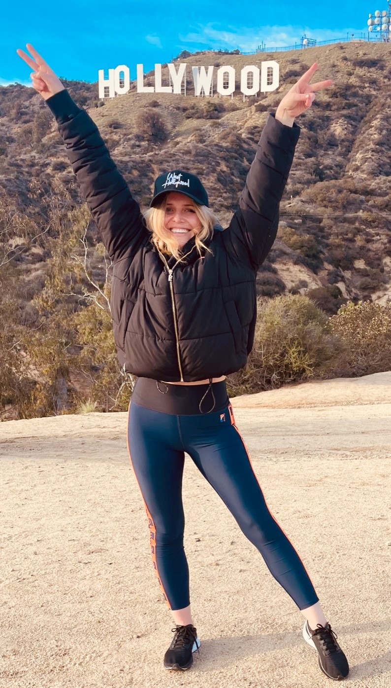 A girl poses with her arms up in front of the Hollywood sign in a brown jacket and leggings. A girl poses in front of the Hollywood Sign on our Hollywood Sign Tour