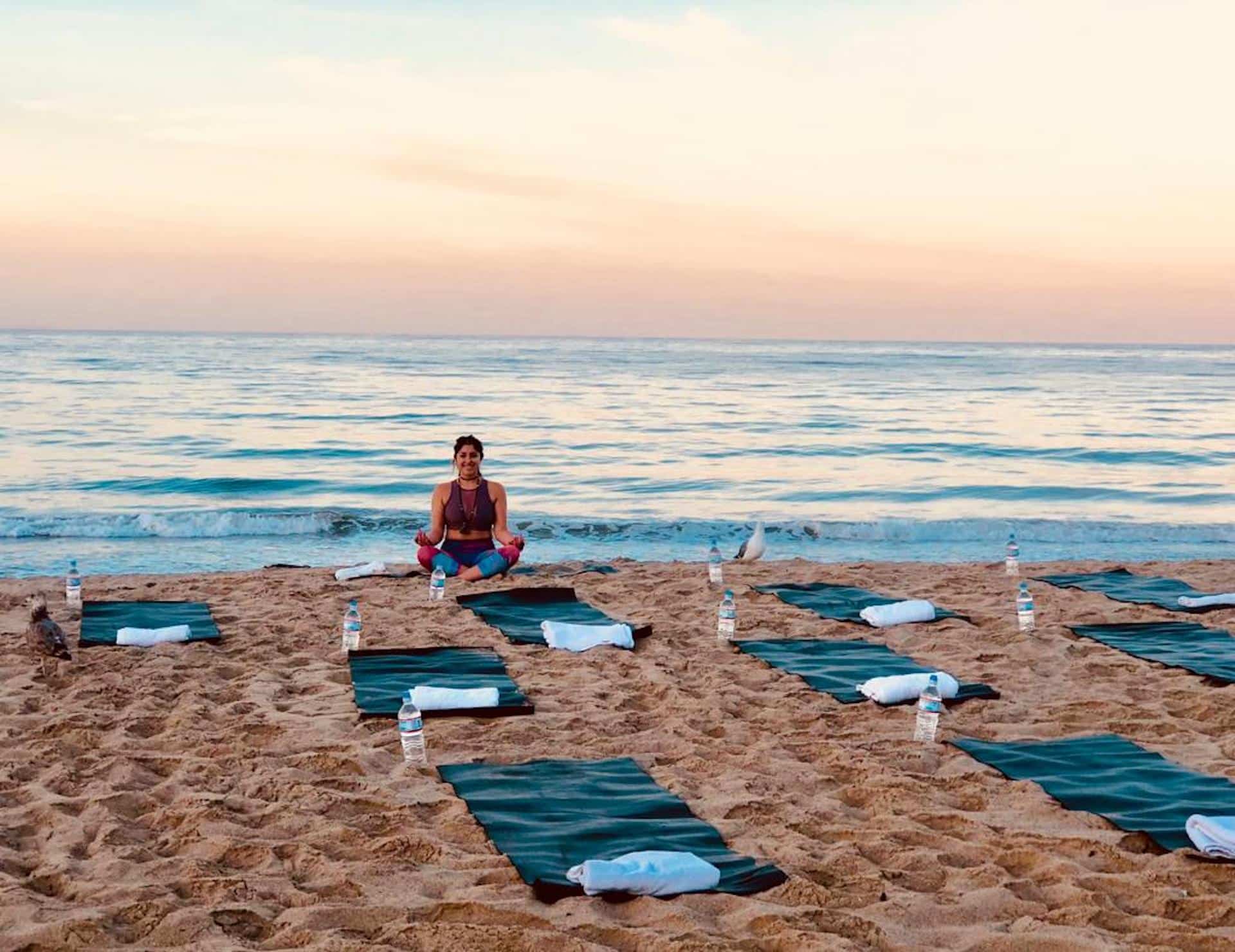 A woman sits on a beach in front of yoga mats at sunset