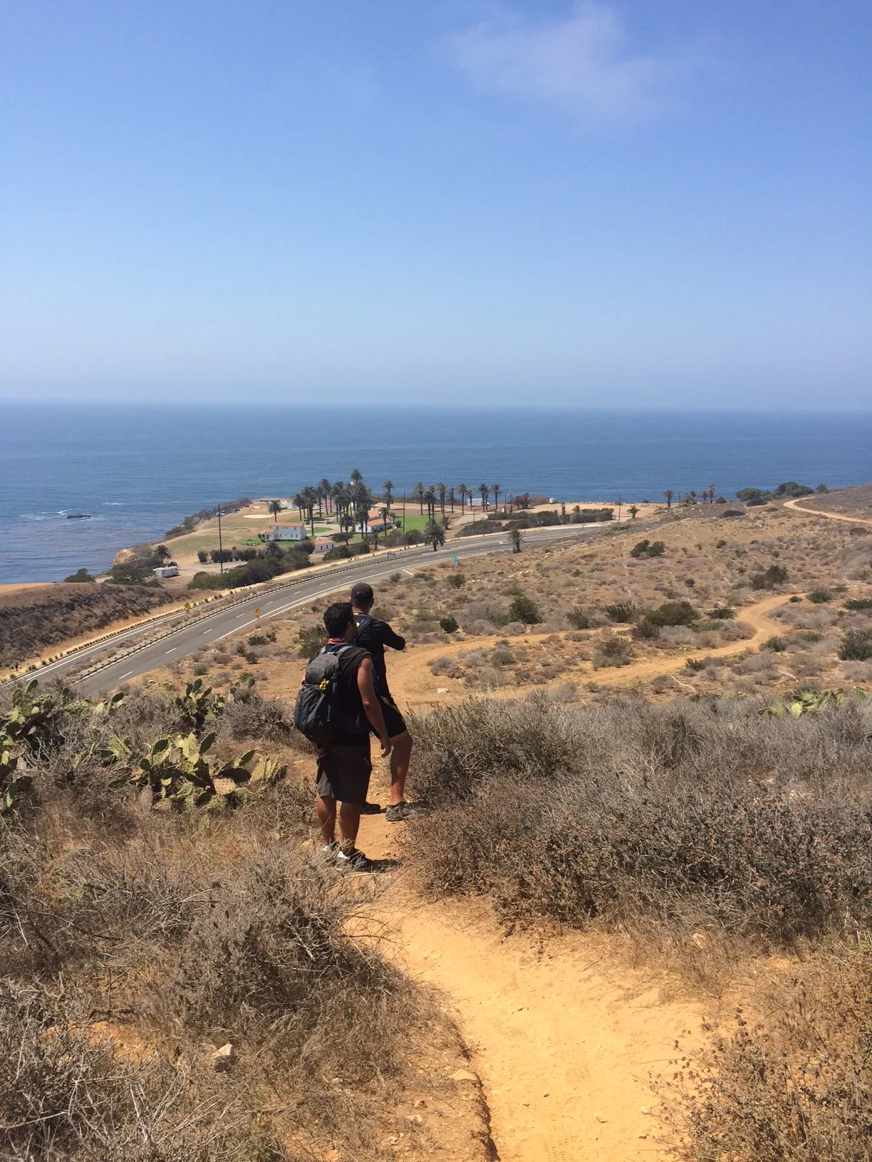 Two hikers standing on a dirt trail overlooking the Rancho Palos Verdes coastline, with ocean views, coastal cliffs, and a winding road below.