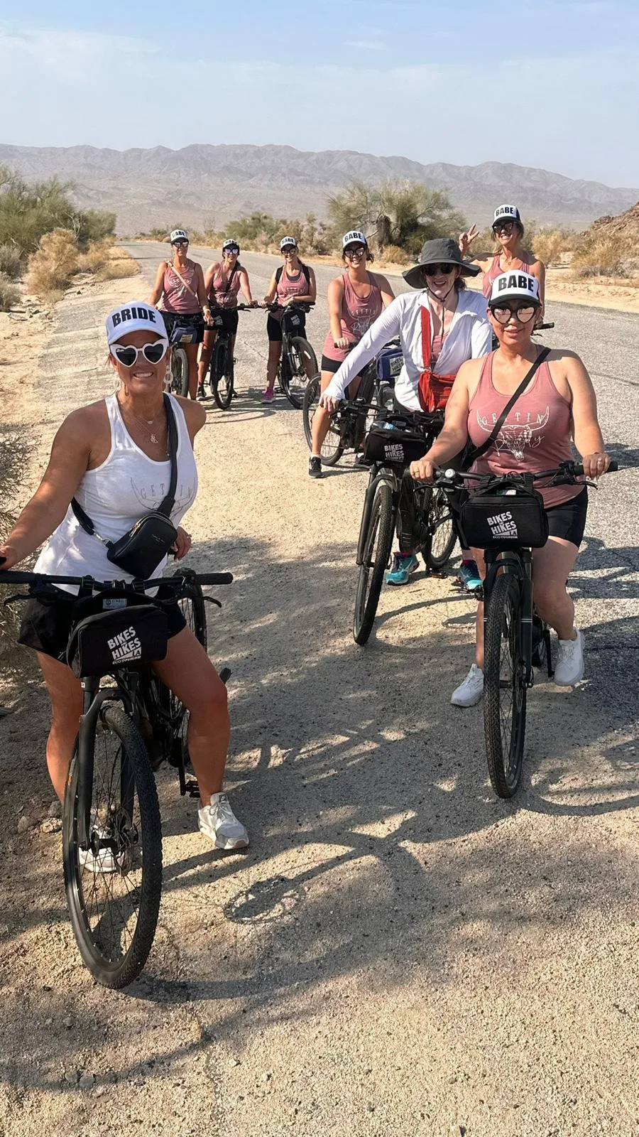 Several women on bikes smile on bikes on a desert road in Joshua Tree