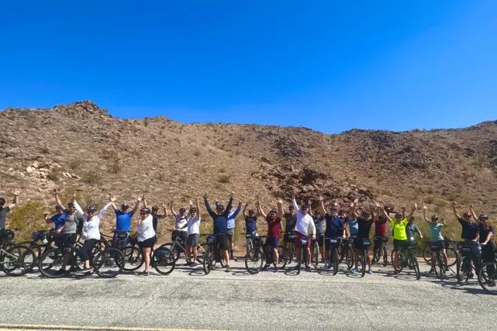 A large group of riders stands in front of a desert hill in Joshua Tree