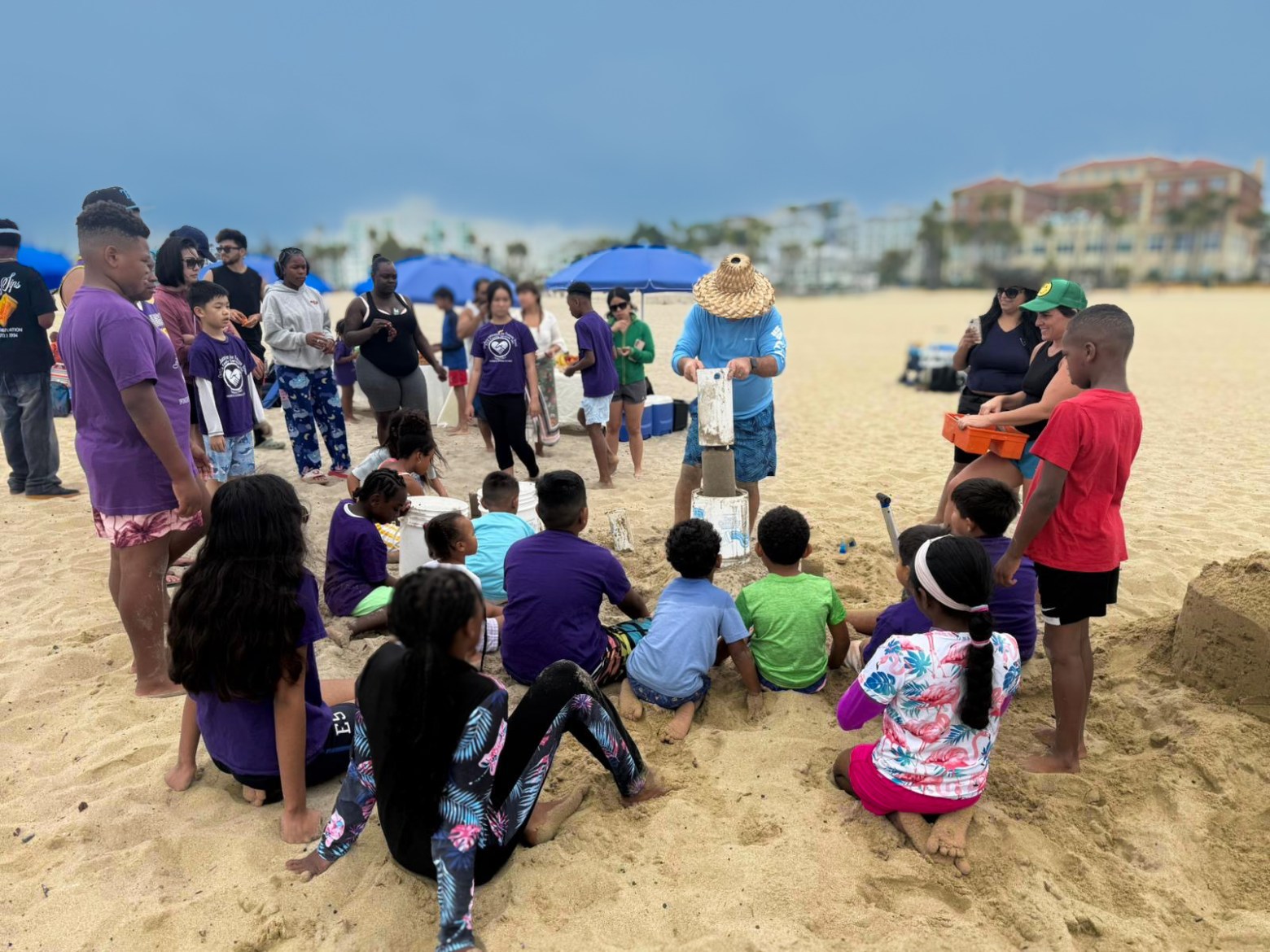 Group of children and adults gathered on a beach watching a demonstration.