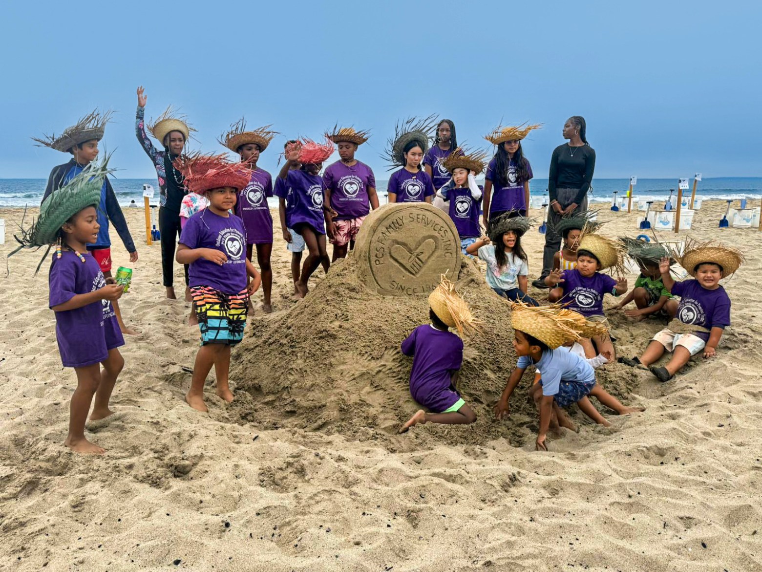 Children in purple shirts with straw hats around a sand sculpture on the beach.