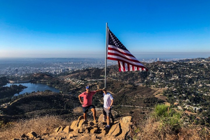 Two hikers stand under an American Flag by the Hollywood Sign