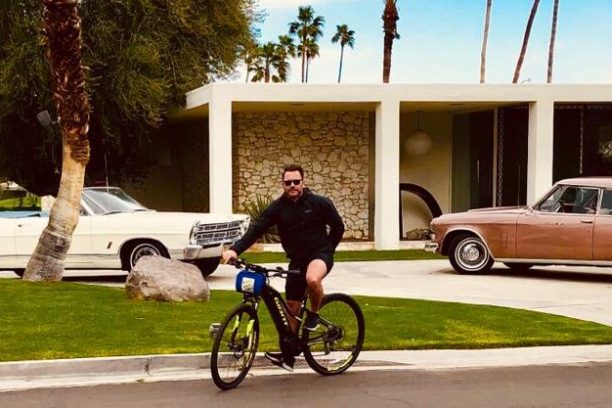 a guest stands in front of a white house in palm springs with a bike on a group tour