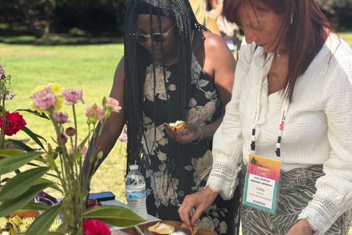 Two women serving food from an outdoor buffet table under a fringed umbrella.