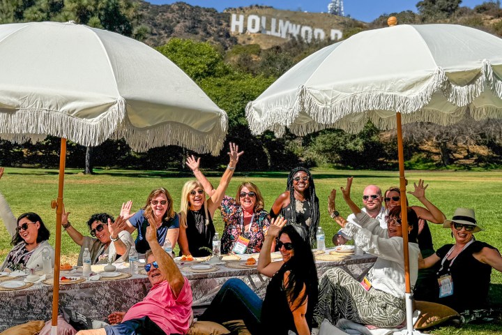 Group of people sitting outdoors under umbrellas, with the Hollywood sign in the background.